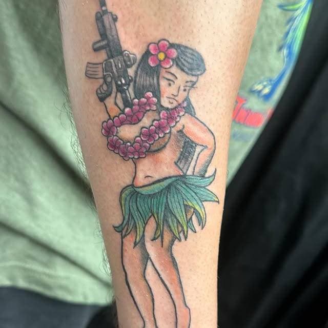 Cartoon hula girl with flower necklace and gun tattoo on forearm.