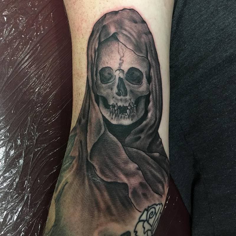 A tattoo of a hooded skull on the forearm in black and grey.
