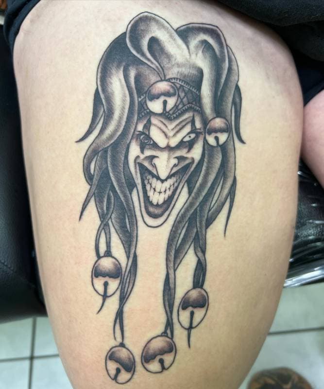 Tattoo of a menacing jester face in black and grey on a thigh.
